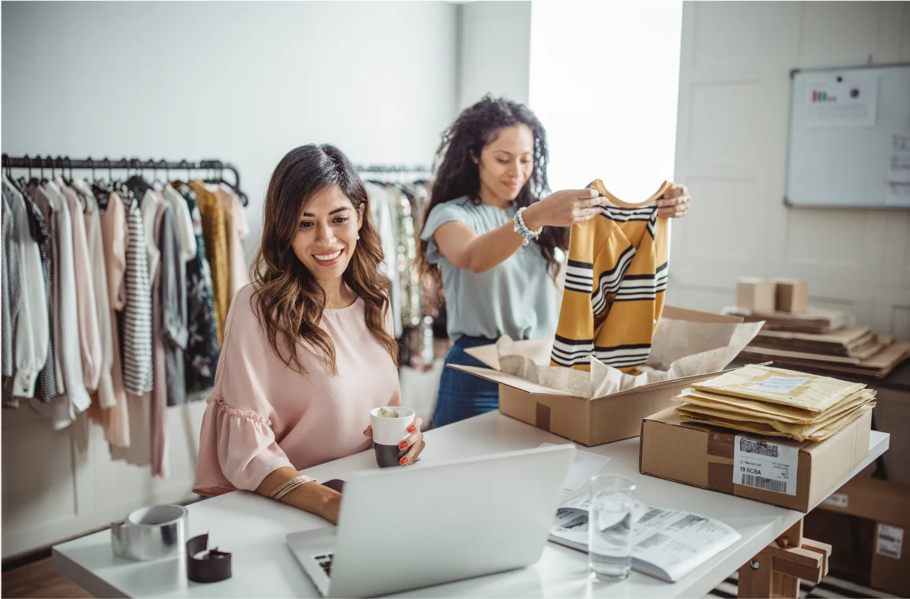 2 women at a desk, one is packaging a t-shirt into a box and the other is on a laptop